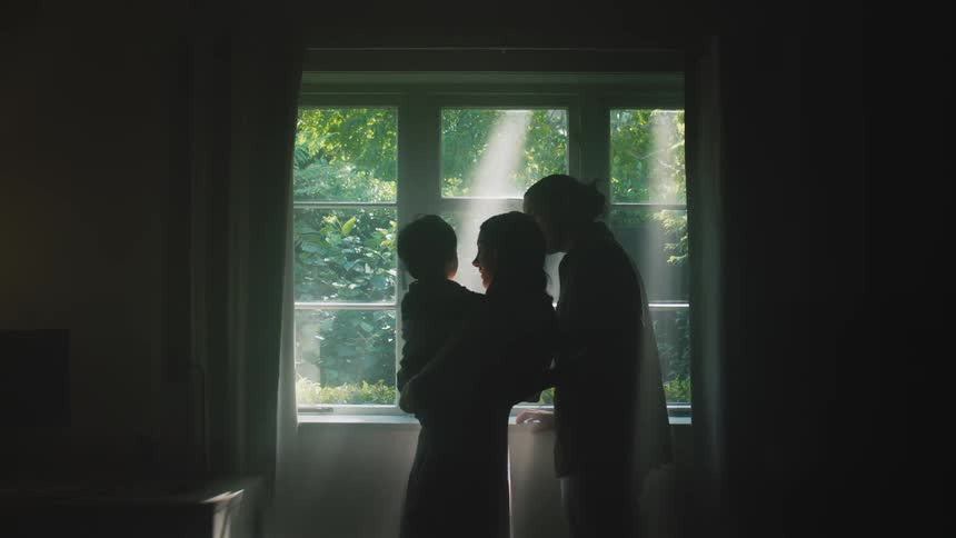 A couple stand in front of a window in silhouette as the mother holds her baby son
