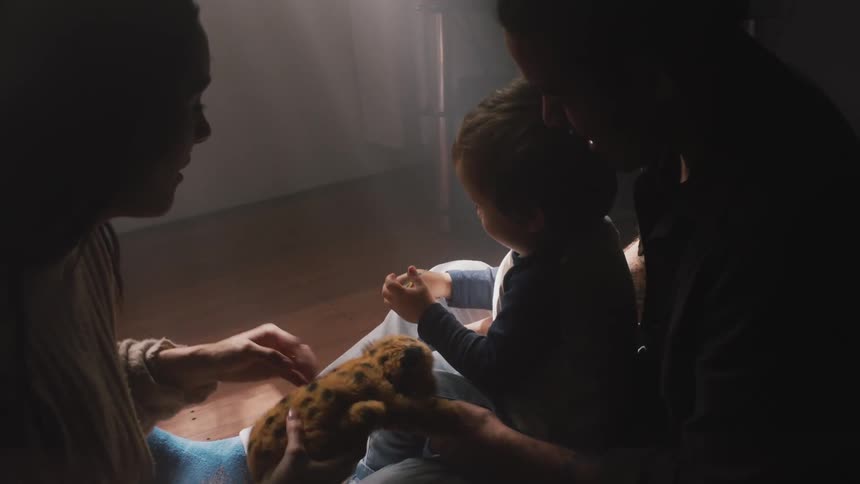 Two women and a toddler playing with a toy on the floor of a dimly lit room at home