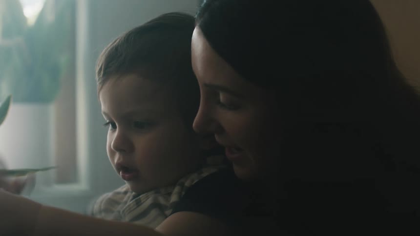 A woman is holding her young son in her arms as he unscrews the top of a milk bottle