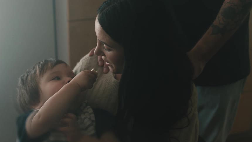A woman is holding her young son in her arms as he reaches his hand up in the air to share his cookie and someone is standing behind her with their hand on her shoulder
