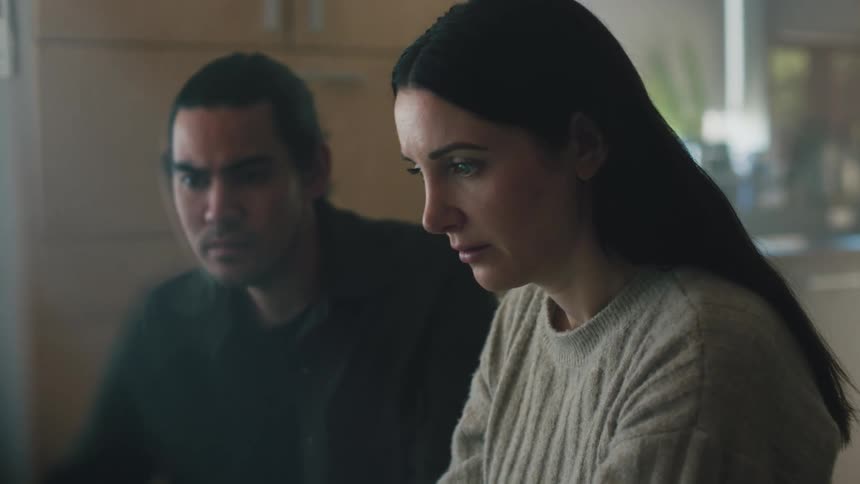 A woman is sitting at a table looking ahead and a man is sitting next to her watching