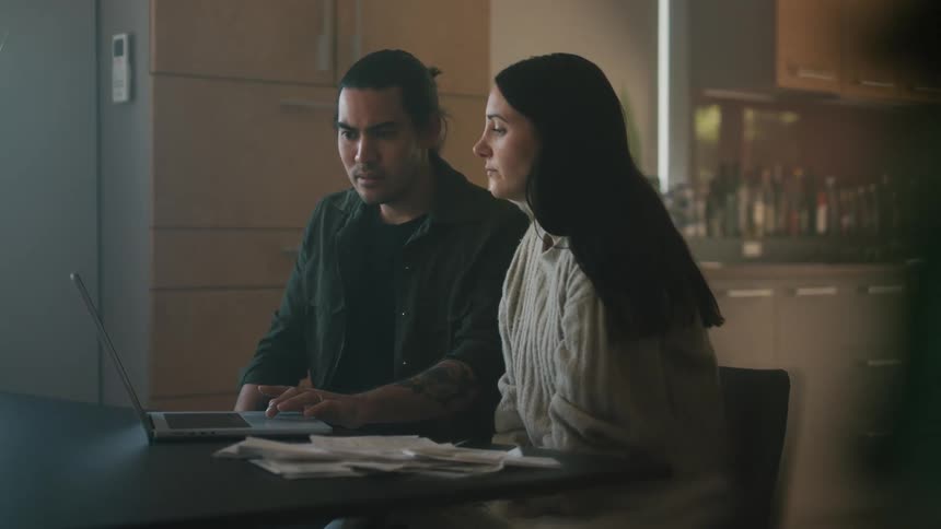 Man in black shirt and woman with straight hair and sweater sit at table and use laptop in the kitchen