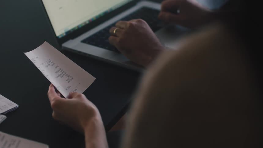 A woman holds a receipt while a man types on a laptop computer