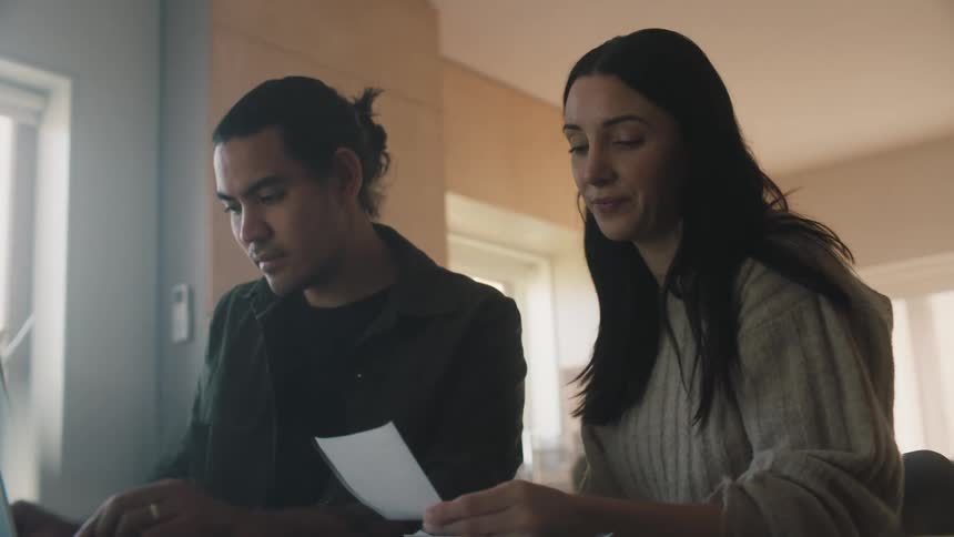 Mother with gray sweater dark straight hair reads paper while man with dark jacket types on laptop in room