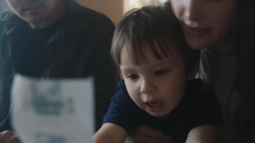A woman is holding her young son on her lap as he looks at a piece of paper and there is a man sitting on the couch next to them