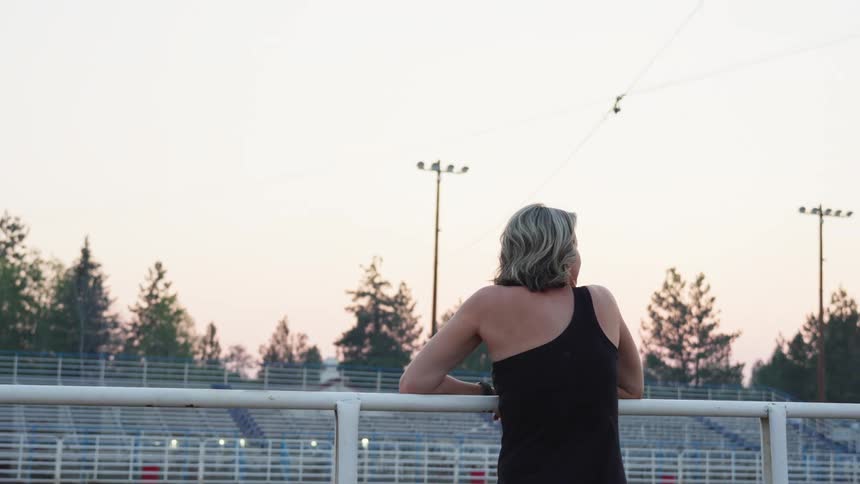 Blonde woman climbs onto rail across from audience stands and looks over arena grounds