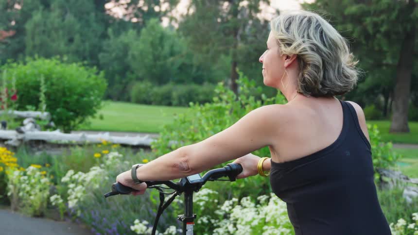 A woman with short wavy hair riding a bicycle in a lush flower filled park