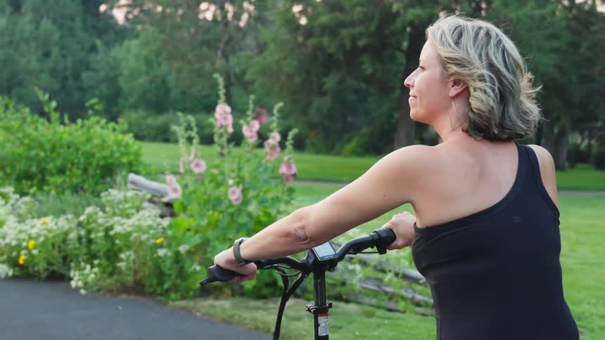 A blonde woman is riding a bicycle and beside her there are plants and green vegetation