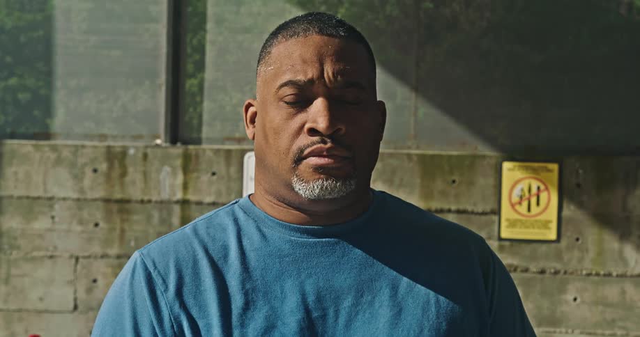 A black man with a gray beard stands outside near a sign on a brick wall