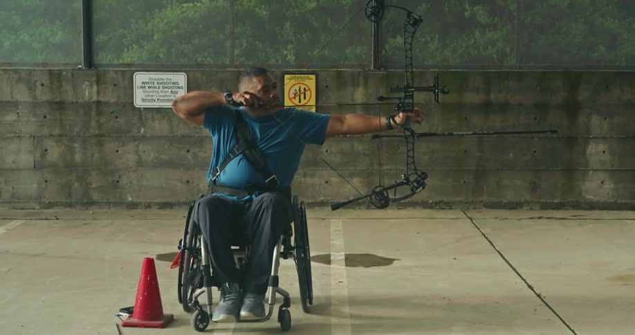 A man in a wheelchair using a professional string bow to aim an arrow then letting the arrow fly