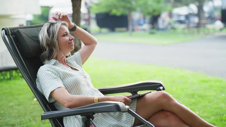 A smiling white woman sitting outside in a suburb closes her laptop and reclines in a chair