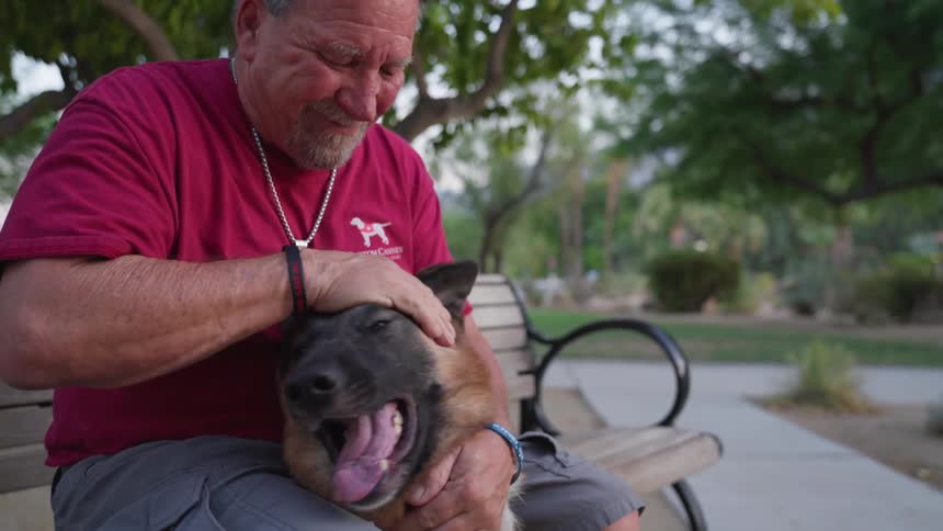 A smiling man sitting on a park bench pets his german shepherd