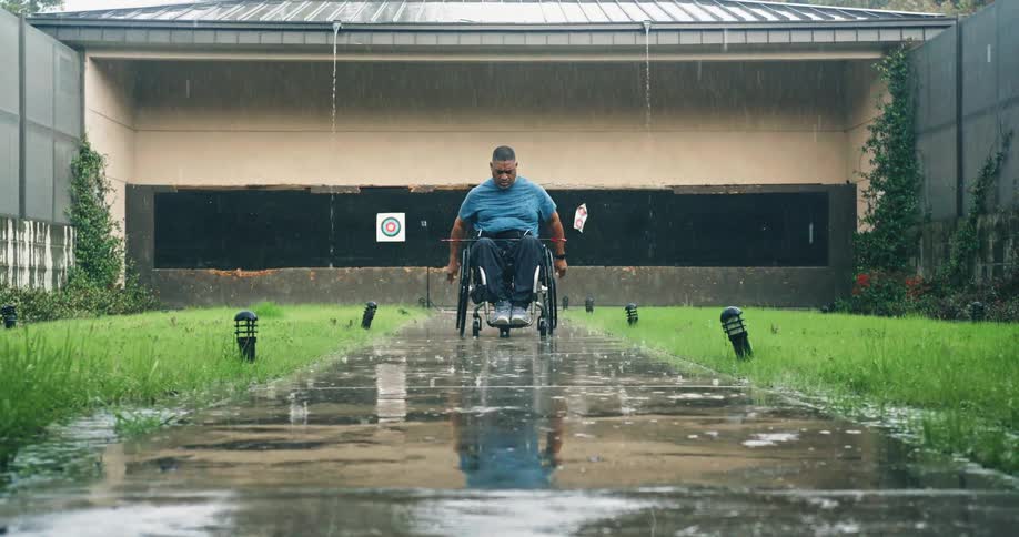 A man in a wheelchair wheels himself away from a shooting range through a concrete path across a lawn