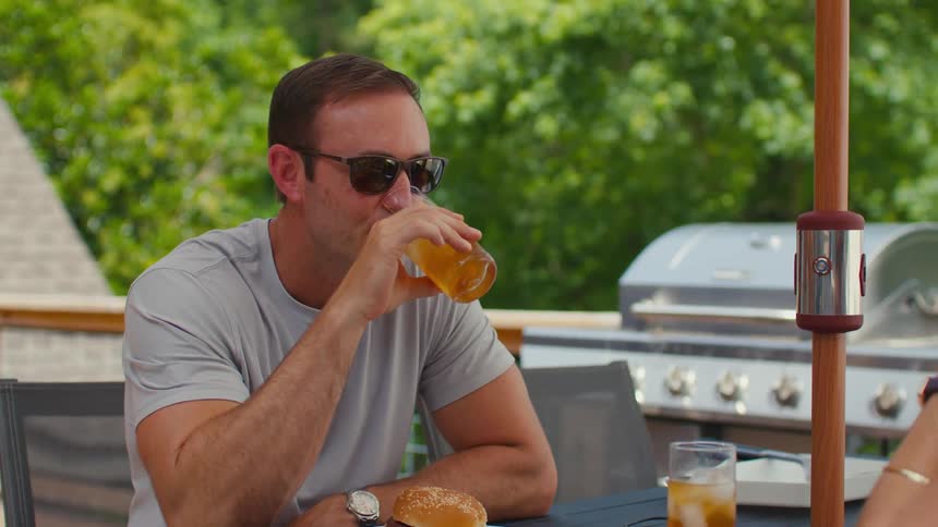A man listens to another person talking while he drinks an iced tea on a deck