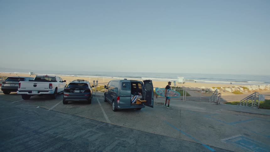 A beachside parking lot with several vehicles including a van with open back doors and people taking surfboards to the beach