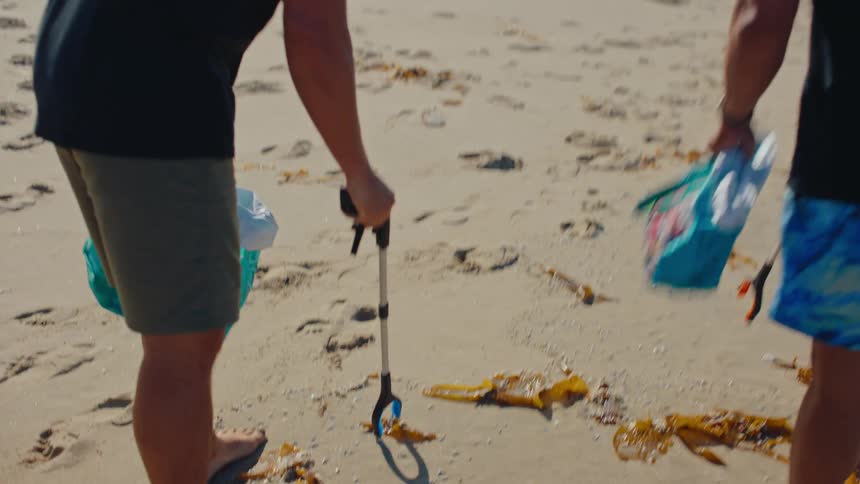 Two men walking on a beach using plastic pincers to collect garbage from the sand into a bag