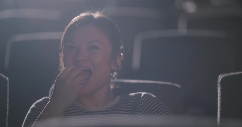 A young woman eats popcorn and laughs while watching a movie in a theater