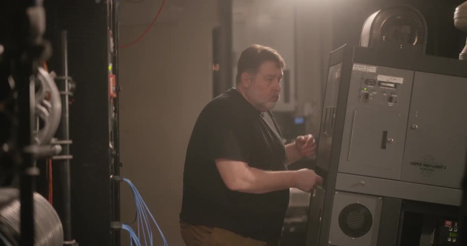 The projectionist is working in the projection room adjusting the projector and inspecting the machinery