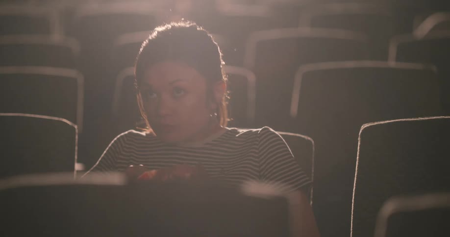 A woman holding a bucket of popcorn is sitting in a movie theater chair with a bright light in the background