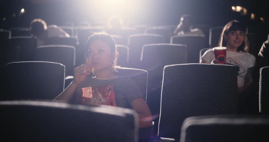 Some people sitting in movie theater chairs while eating popcorn staring straight ahead and there is a bright light behind them