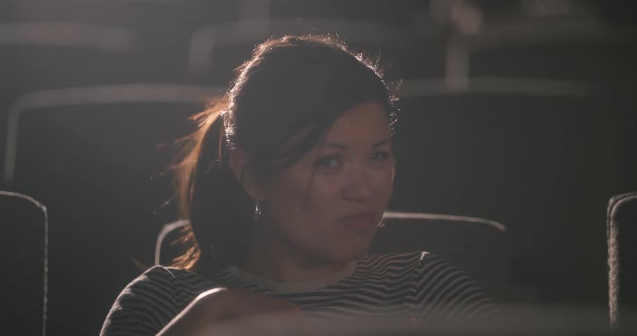A closeup of a womans face in a movie theater her hands covering her face as she turns away in fear