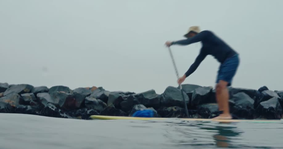 A man rows a waterboard across the water near a pile of rocks