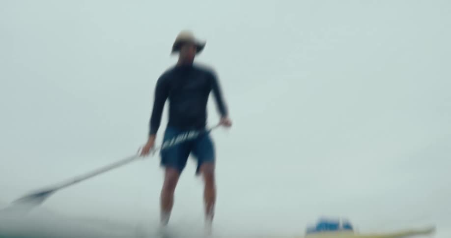 A man standing on a surfboard using an oar to ride a wave towards the beach
