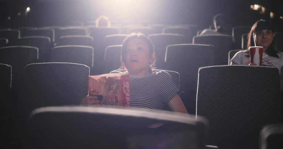 A woman tosses her popcorn in fright as another viewer smiles inside of a movie theater