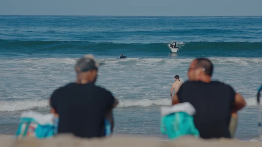 Two men sit on a beach talking while watching surfers at a beach