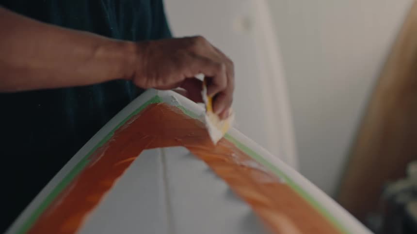 Hand with a spatula applying a white cream sealant to the tip of a surfboard