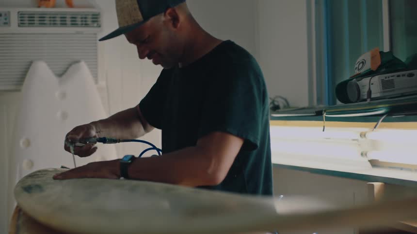 An individual repairs a surfboard using an aircompressed tool while it rests on supports