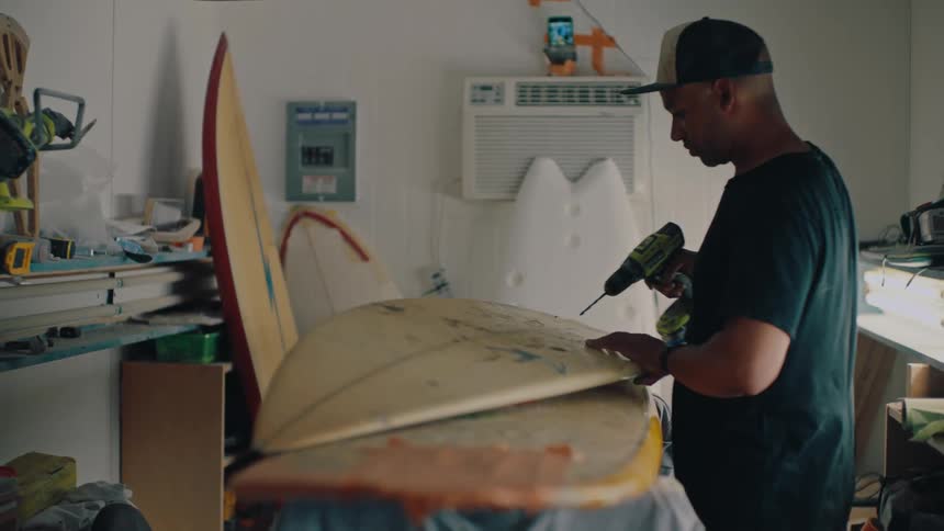 An individual repairs a surfboard using a drill while it rests on supports