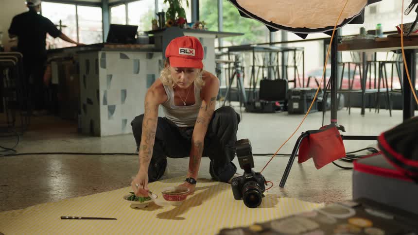 Two women arranging table items and food on the floor for a photoshoot then one of them gets up and walks away