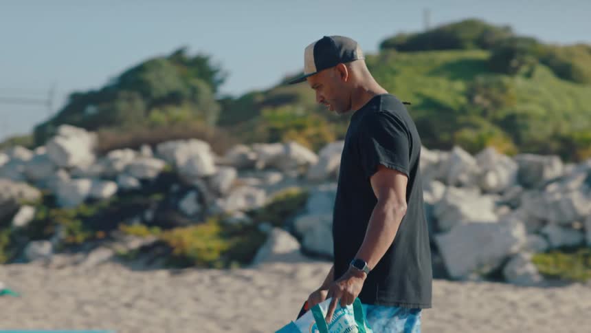 A man in a cap is walking on the sand of a beach and collecting something from the ground using a picker and putting it in a bag