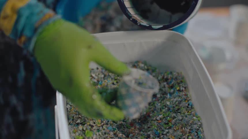 A human hand wearing a yellow glove taking some microplastic from a jar and placing it into a smaller one