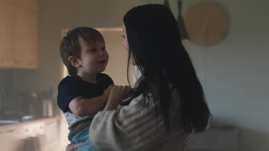 A mother holding a young child in her arms in a kitchen with soft lighting