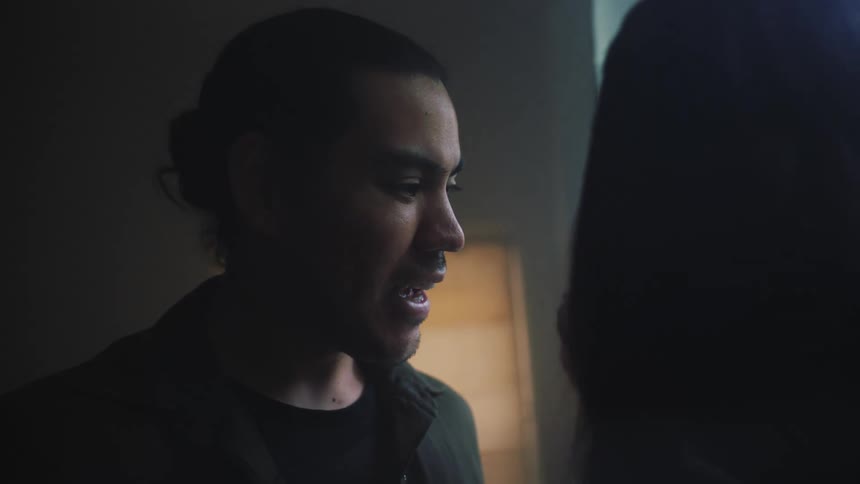 Close up of the face of a young person with dark hair tied back talking to someone