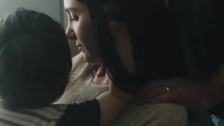 Mans hands resting on the shoulders of a woman who is holding a small child and giving kisses