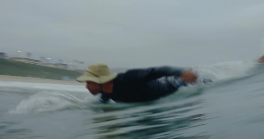 A man wearing a hat is paddling on his board to catch a wave