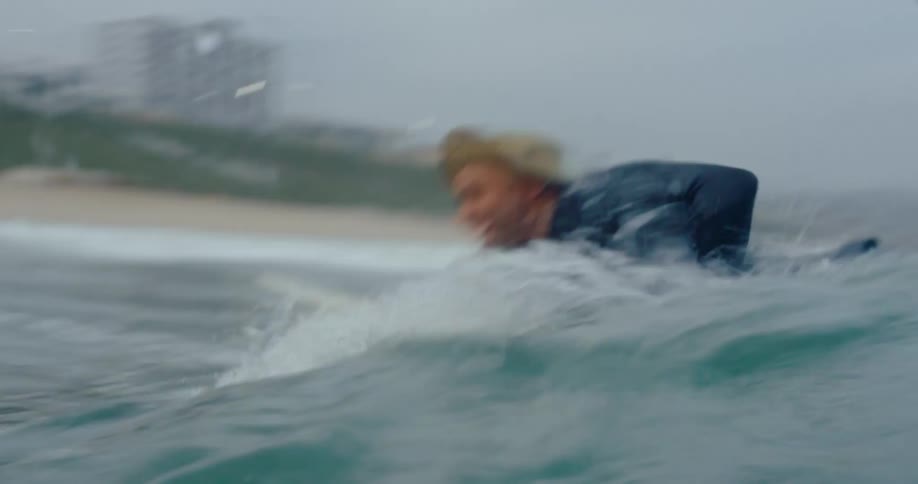 A man wearing a hat is lying on a surfboard and paddling to catch a wave