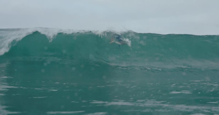 Wave breaks over the head of the surfer riding it on a cloudy day