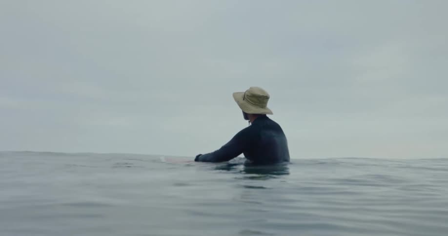 A person in a wetsuit and a sun hat rides a surfboard in the ocean