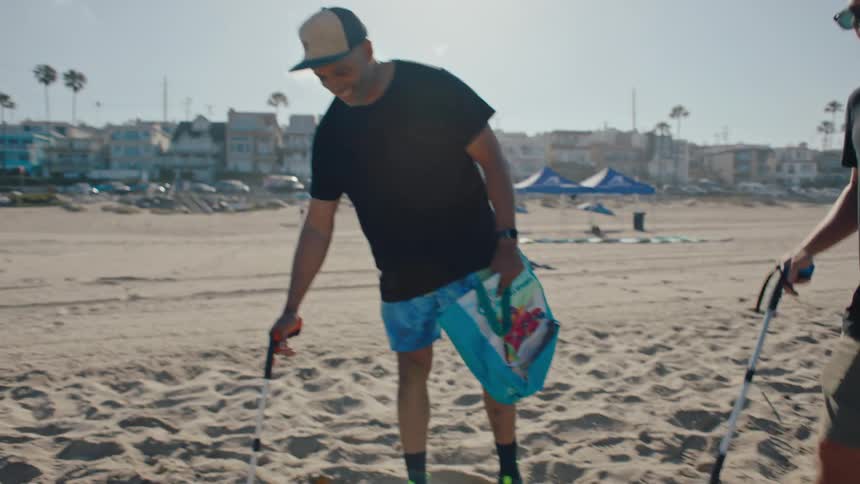 Two men with tongs and bags are cleaning sand from a beach as they walk