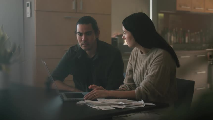 A worried looking woman taps on a laptop while talking to man in a dining room
