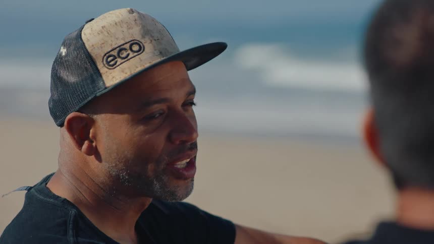 A man wearing a cap is talking to someone in front of him and they are on a beach