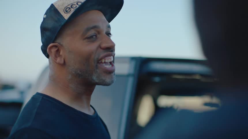 A man wearing a cap is talking to someone on his right and he is smiling as he speaks