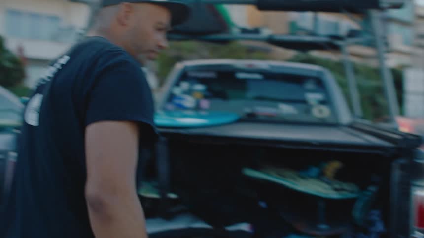 A man wearing sunglasses approaches another man wearing a cap and he places his surfboard in the back of an open pickup truck