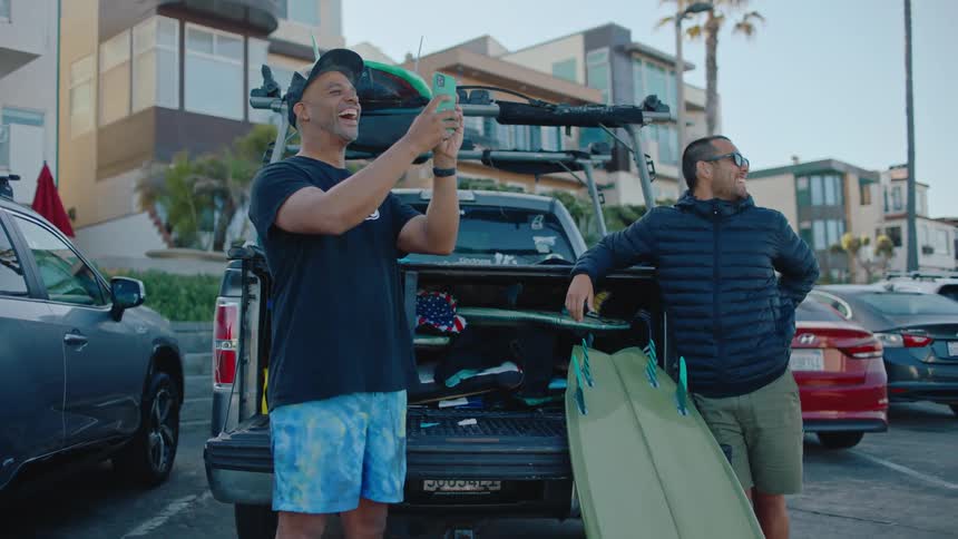 A man leaning on a truck with a board sticking out and another man has his cell phone pointed forward and then he looks to the side and raises his arms up