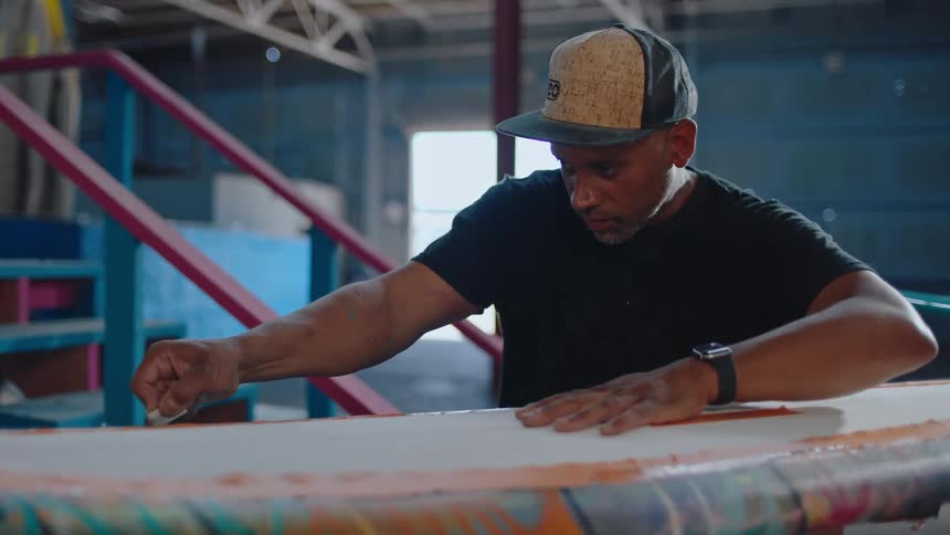A man in a cap leaning over a large colorful surfboard cutting the surface with a razor