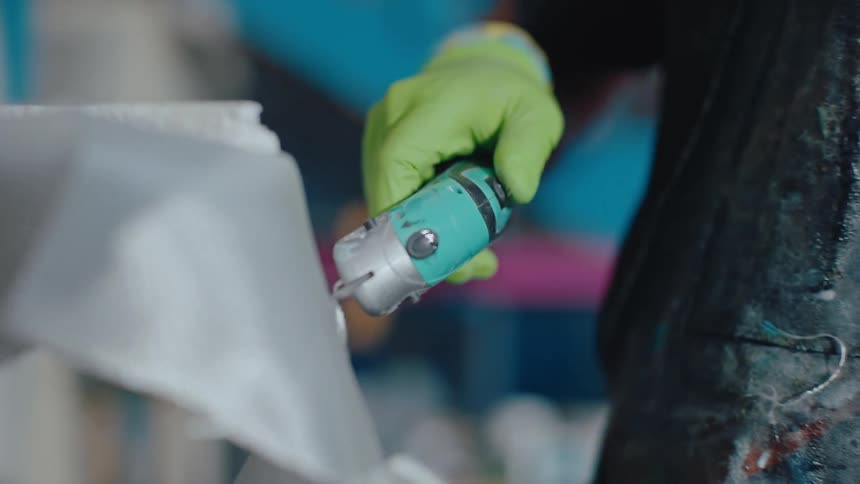 Closeup of a hand wearing a green glove cutting fabric with an electric tool